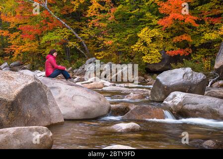 Lonely thoughtful woman sitting on a rock on a mountain river on a fall day. Beautiful autumn colours in background. Stock Photo