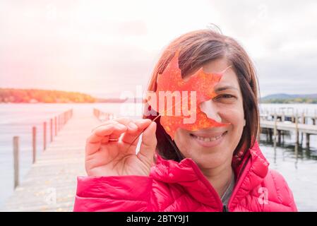 Portrain of a smiling woman holding a red maple leaf in front of her right eye. A deserted jetty on a lake is visible in background. Stock Photo