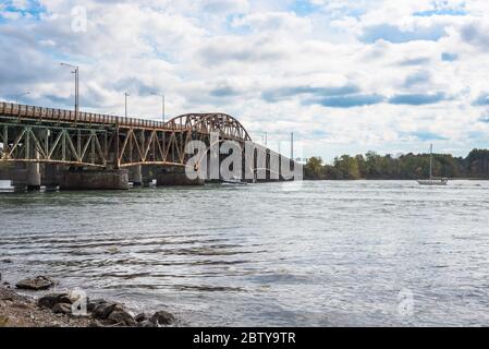 Boats sailing up a mighty river passing an old rusty steel road bridge on a cloudy autumn day Stock Photo