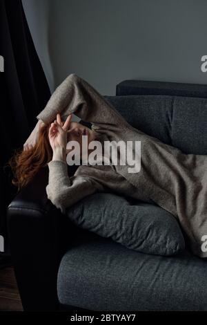 The red-haired girl lies on the sofa, closing her eyes with her hand, shows the peace sign with her fingers. Stock Photo
