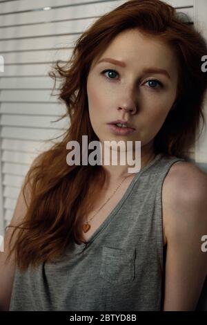 A beautiful redhead girl with her hair and freckles in a gray T-shirt is looking at the camera, leaning on a white wooden screen Stock Photo