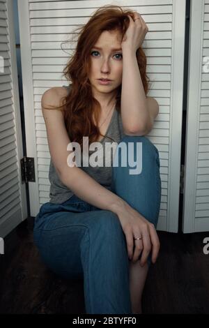 Redhead girl with freckles in a gray T-shirt is looking at the camera, sitting on the floor and leaning on a white wooden screen. Stock Photo