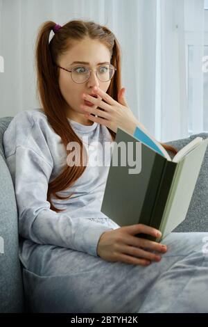 A red-haired girl in glasses and pajamas looks into the book shocked by opening her eyes and covering her mouth with her hand Stock Photo