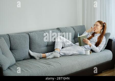 A red-haired girl in pajamas and glasses lies on the couch with a book and yawns Stock Photo