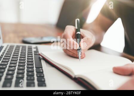 close-up of person taking notes with pen on paper in front of laptop computer Stock Photo