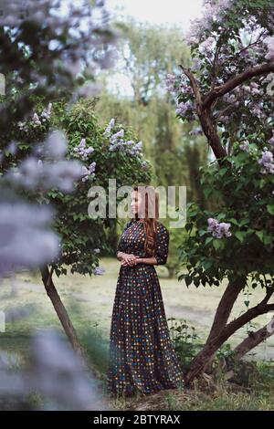 Beautiful dreaming redhead young woman posing in a long dress by a blooming lilac in the spring Stock Photo