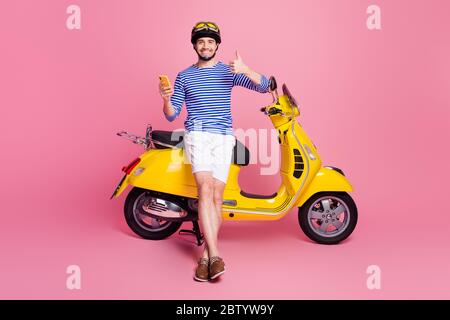 Portrait of his he nice attractive handsome cheerful cheery guy sitting on moped using cell app 5g showing thumbup advert ad cool isolated over pink Stock Photo