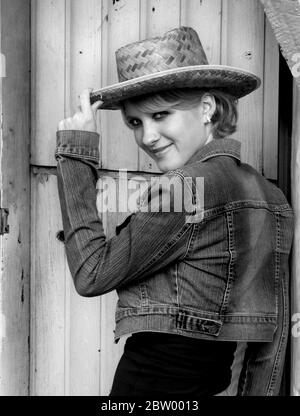 Black and White closeup of young woman wearing denim jeans and jacket.  She is tipping the brim of her straw hat and grinning. Stock Photo