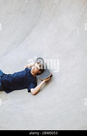 Cool young guy thoughtfully looking in camera while lying on skateboard in empty bowl at modern skatepark Stock Photo