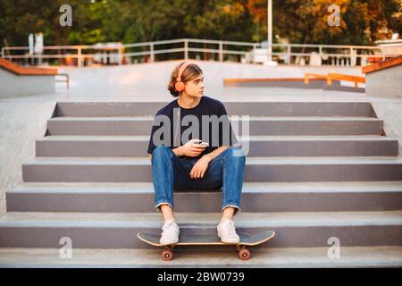 Young skater in orange headphones with cellphone in hands sitting on stairs at skate park Stock Photo