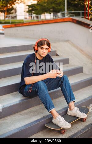 Young skater in orange headphones holding cellphone in hands thoughtfully looking in camera sitting on stairs at skate park Stock Photo