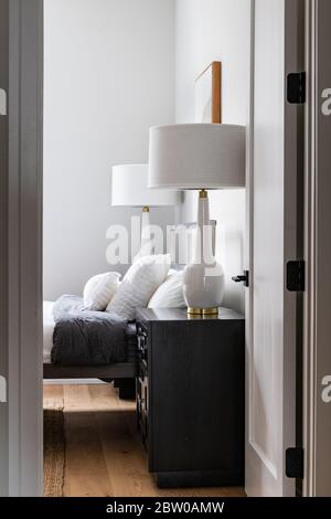 Looking into a modern bedroom through the doorway. White lights sit on nightstands around a high bed with white bedding. Stock Photo