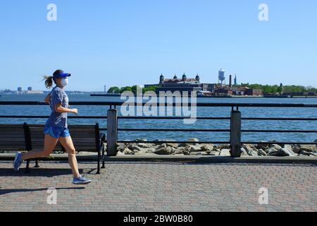 A female jogger wearing mask jogging on Hudson River Waterfront Walkway with Ellis Island in the background.Liberty State Park.Jersey City.New Jersey.USA Stock Photo