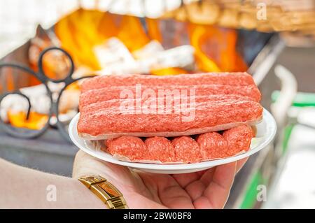 Chevapchichi on a plate in your hand. A man holds fresh chevapchichi on a white platter against the background of a barbecue fire. Stock Photo