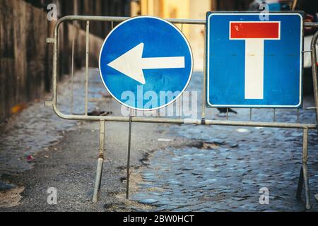 Road works detour road signs. Dead end road and arrow to turn left. Iron crossing barrier. Street with paving cobblestones. Stock Photo