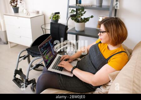 Serious disabled woman in eyeglasses sitting on sofa and using laptop for coding at home Stock Photo