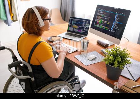 Female freelance programmer in modern headphones sitting in wheelchair and using computers while coding web game at home Stock Photo