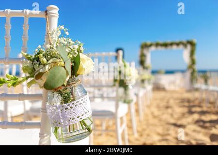Luxurious wedding ceremony on the ocean, beach. White chairs decorated with a beautiful bouquet of flowers in a jar hanging on them.  Stock Photo