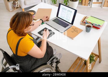 Disabled UI designer sitting in wheelchair and viewing sketch on laptop while working on interface Stock Photo
