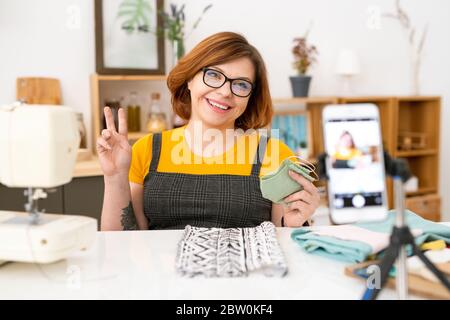 Cheerful young seamstress in eyeglasses sitting at desk with fabrics and holding cloth mask while shooting video lesson at home Stock Photo