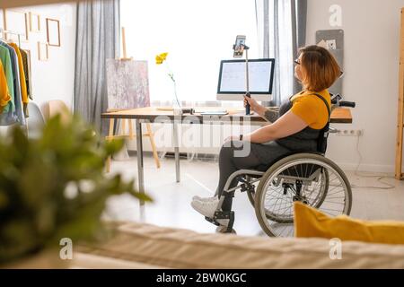 Disabled young woman sitting in wheelchair and using monopod while taking selfie at home Stock Photo
