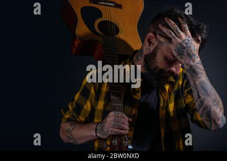 Tattooed man with a beard wearing a tartan yellow black shirt is  posing holding his guitar on his sholder Stock Photo