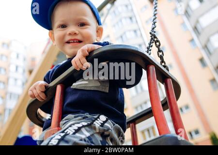 Funny and handsome boy rejoices, rides on a children's swing, plays on the playground Stock Photo