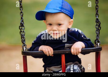 Funny and handsome boy rejoices, rides on a children's swing, plays on the playground, 1-2 years Stock Photo