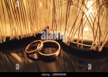 Pair of wedding rings laying on dark reflective surface in front of decorative light. Toned to warm colors. Wedding day concept. Stock Photo