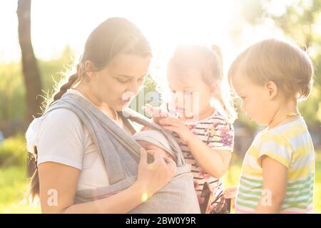Motherhood, care, infants, summer, childhood and large families concept - Young beautiful mom with newborn baby in sling and two small children of the Stock Photo