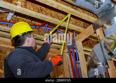 Male builder measuring plastic pipes with new home construction in hot and cold blue and red pex pipe layout in pipes exposed beams Stock Photo