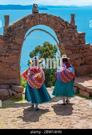 Two Peruvian Quechua indigenous women in traditional clothes walking through the arch of the rulers on Taquile island with the Titicaca Lake, Peru. Stock Photo