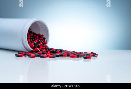 Red-black antibiotic capsule pills spread out of white plastic drug bottle on gradient background. Pharmaceutical industry. Antibiotic drug resistance Stock Photo