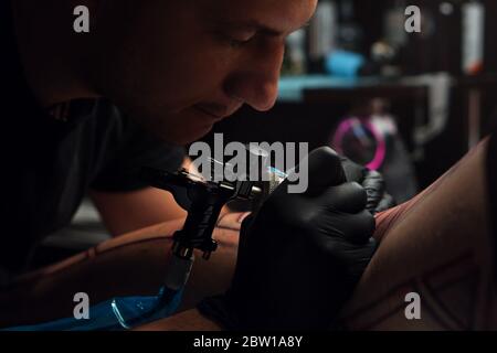 Process of making tattoo close-up. Professional tattoo artist makes tattoo on young man leg. Master doing tattoo in salon.  Stock Photo