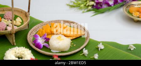 Cropped shot of several Thai traditional dessert serving on wooden tray with flower and banana leaves decorated on white table Stock Photo