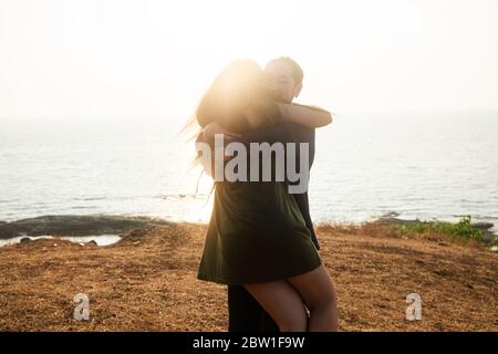 Backlit Portrait of a young romantic couple swirls in a hug against the backdrop of the sea. Anjuna Hills, Goa, India Stock Photo