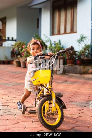 Margao, Goa/India - May 10 2020 : Playful Pretty Indian girl child/infant/toddler wearing a hair band, playing with a cycle/tricycle. Stock Photo