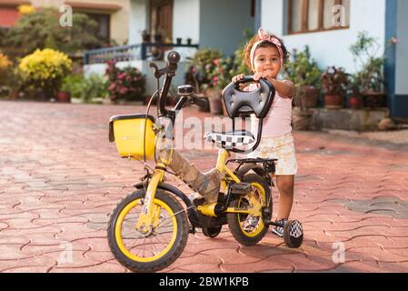 Margao, Goa/India - May 10 2020 : Playful Pretty Indian girl child/infant/toddler wearing a hair band, playing with a cycle/tricycle. Stock Photo