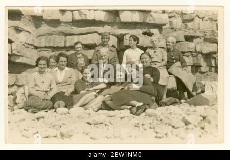 Early 1900's postcard of large group of older people, old-fashioned clothes, frumpy, day-trippers, sitting on a on beach in the shelter of a rocky cliff, circa 1930's, U.K. Retro beach photo. Stock Photo