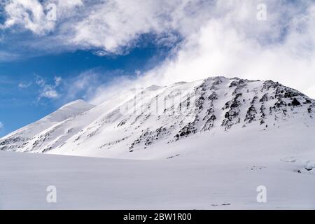 Big mountain in a snowy landscape against a blue sky in front of a smooth snow covered ground. Clouds are rising behind the mountain looking like a pi Stock Photo