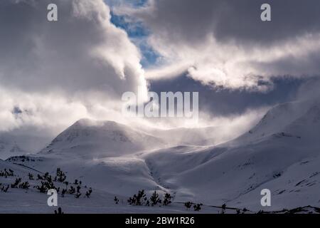 Dramatic clouds gathering over a large snow covered mountain bringing it into its shadow. Sun rays are seen shining through the dark clouds. Stock Photo