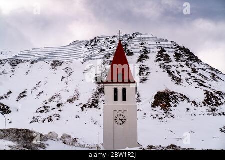 A pointy church with a bright red roof and a cross is towering against the cloudy skies in front of a snow covered mountain. Stock Photo