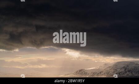 Sun rays shining through the dark clouds after a storm illuminating the large mountains with a golden light. Stock Photo
