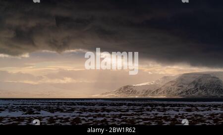 Sun rays shining through the dark clouds after a storm illuminating the large mountains with a golden light. Stock Photo