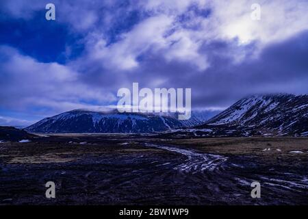 Partly cloud covered snowy mountains and an old muddy road going through the barren landscape Stock Photo