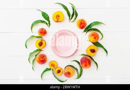 Frame made of peaches and peach tree leaves with pink empty plate in center. Flat lay with copy space. Whole peaches and half peach with stone on Stock Photo