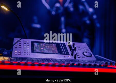 An advanced mixing console in dim coloured lighting, with the stage posing in the background. Stock Photo