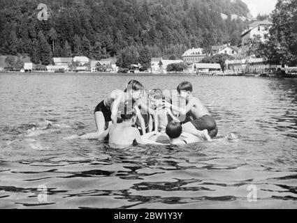 Boy king of Yugoslavia joins in the fun of camp life. King Peter, the boy monarch of Yugoslavia, is spending his summer holiday in camp with a party of Boy Scouts in the grounds of Suvobar, the summer Royal residence of the shores of Lake Bled, King Peter is drawing freely in the life of the camp, helping in the daily tasks and taking part in the games. Like his comrades, he wears only a pair of shorts. Photo shows, King Peter (right) joins in some good natured 'ragging' in the waters of Lake Bled. 14 June 1937 Stock Photo
