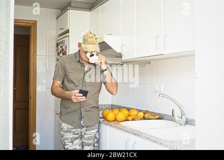 Adult Western male with glasses, cap and activity bracelet drinking espresso in a modern white home kitchen. Green summer clothes. Fruit on the counte Stock Photo
