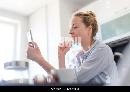 Young pleased woman indoors at home kitchen using social media apps on mobile phone for chatting and stying connected with her loved ones. Stay at Stock Photo